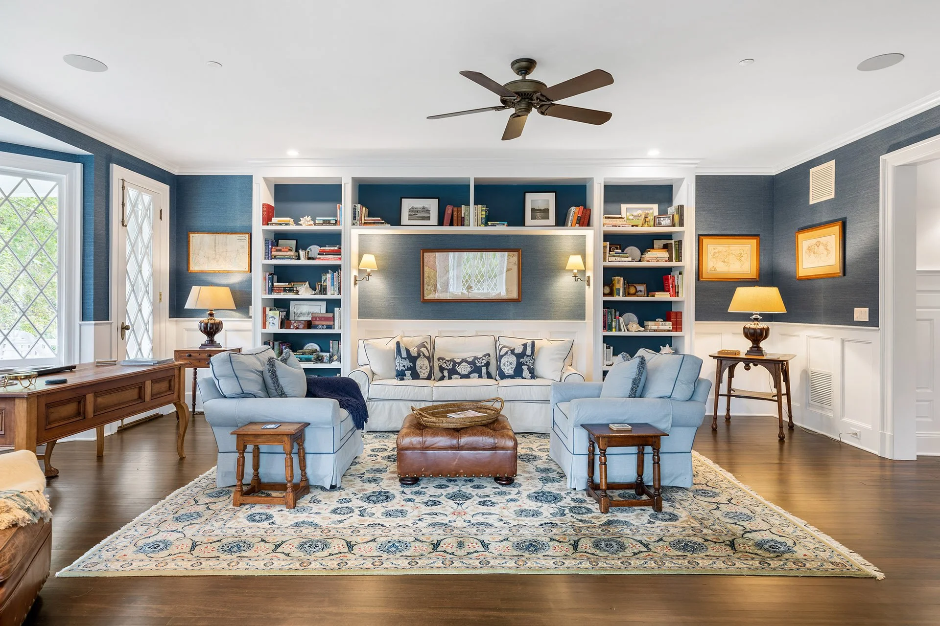 Living room with blue and white walls, white sofas with navy and white pillows, wooden coffee table, built-in shelves with books, framed artwork, and lamps, hardwood floor, and a patterned rug.
