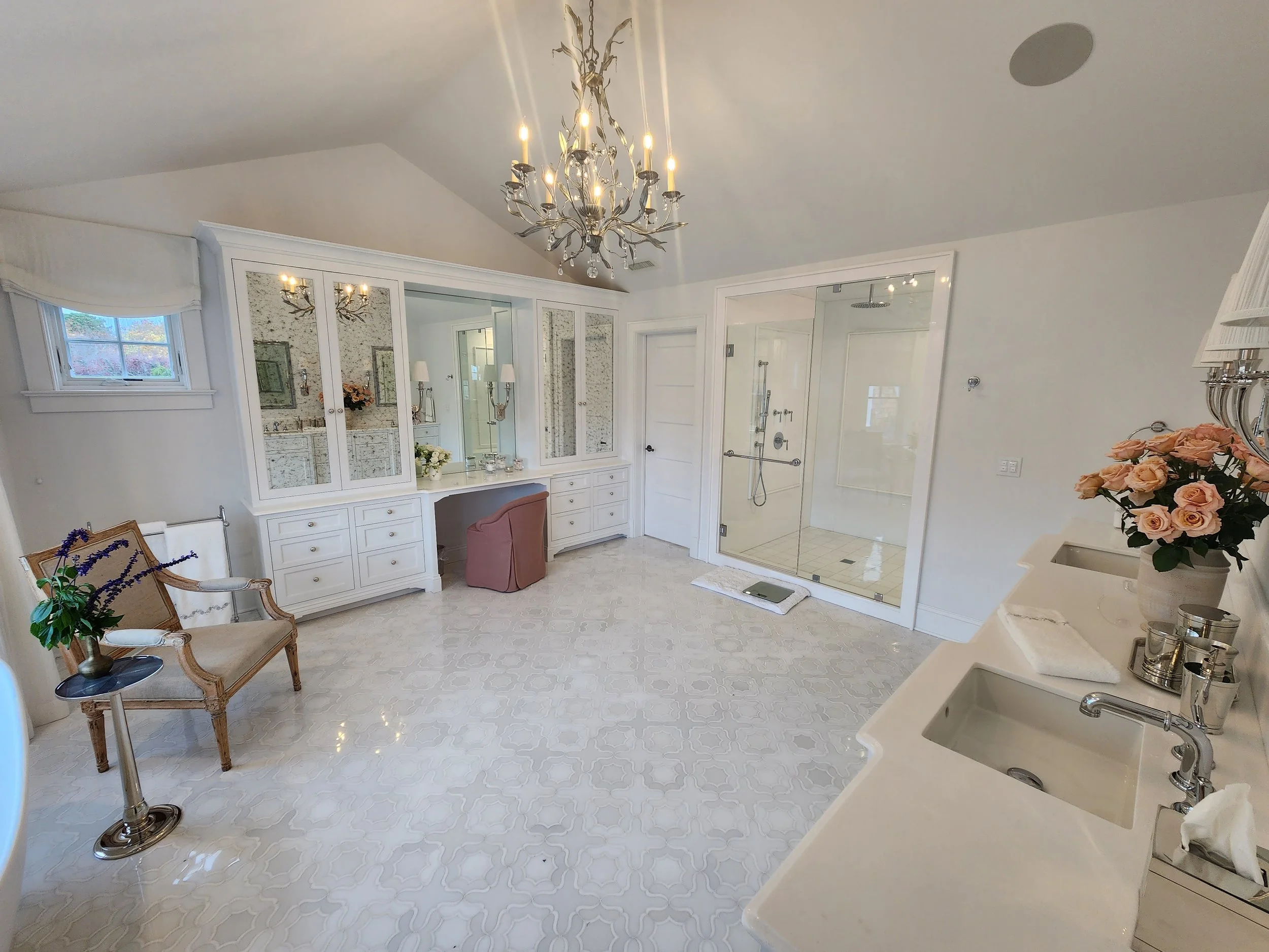 A spacious, well-lit bathroom with a chandelier, a vanity with mirrors and drawers, a glass shower, a vintage-style chair with a pink cushion, a small table with a potted plant, and a sink with a faucet and pink roses.