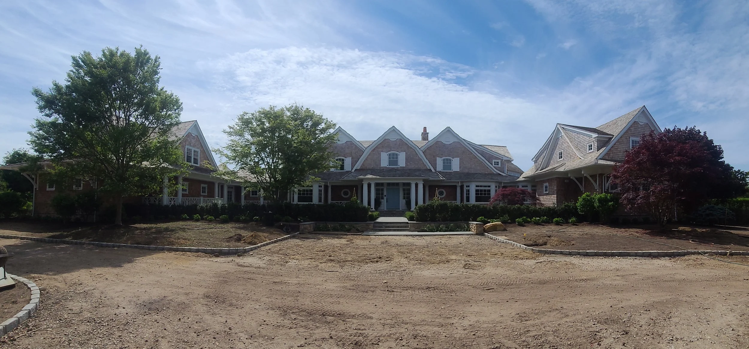 Newly constructed large house with a landscaped front yard, trees, and a dirt driveway under a partly cloudy sky.
