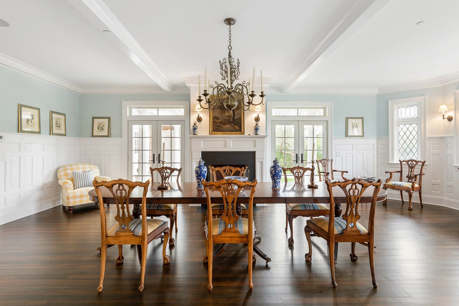 Elegant dining room with a long wooden table, ornate wooden chairs, a chandelier, fireplace, and large windows in a bright, spacious setting.
