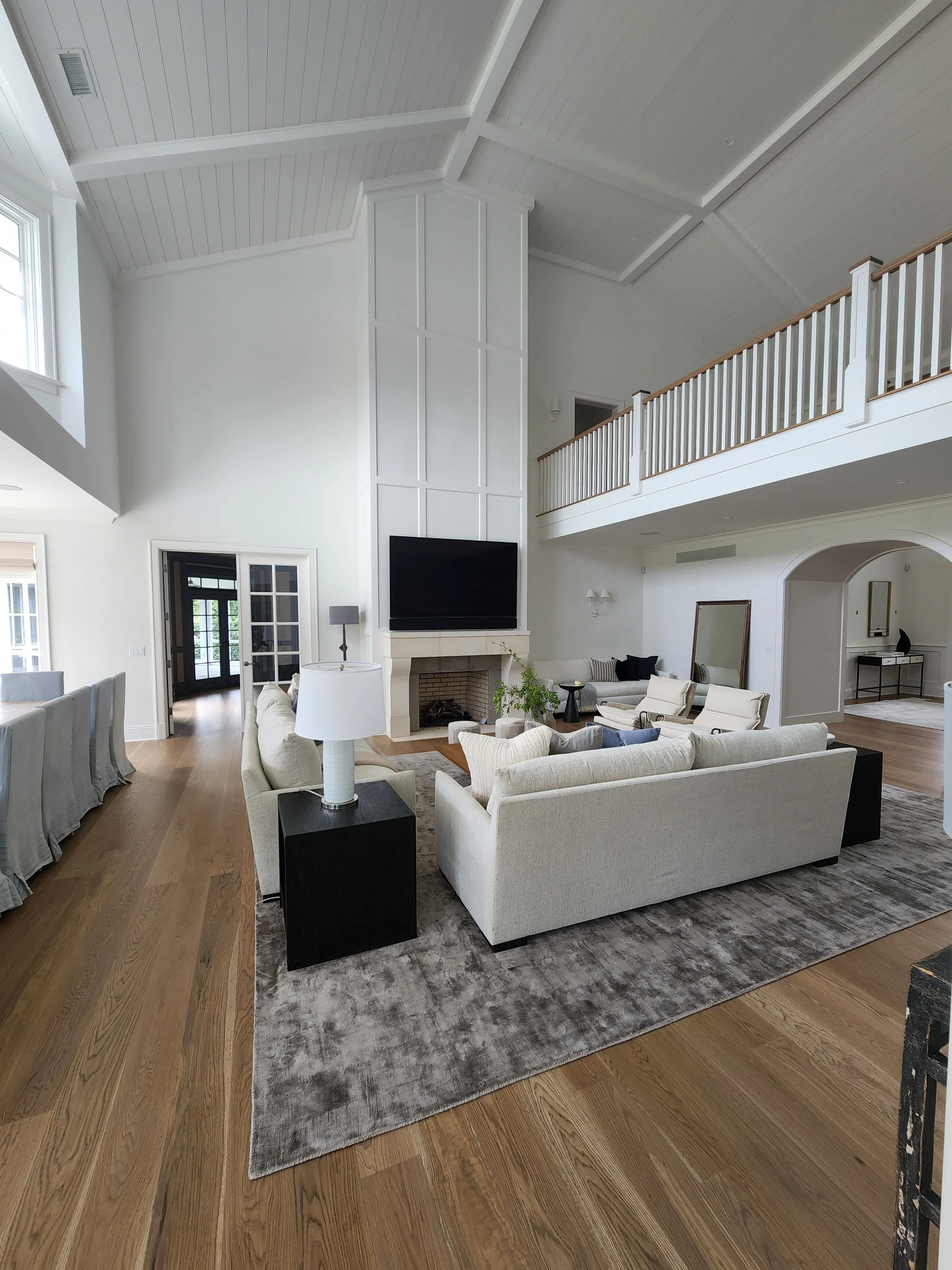 Spacious living room with high white ceiling, white walls, hardwood floors, sectional sofa, armchairs, fireplace with mounted TV, and a balcony railing overlooking the room.
