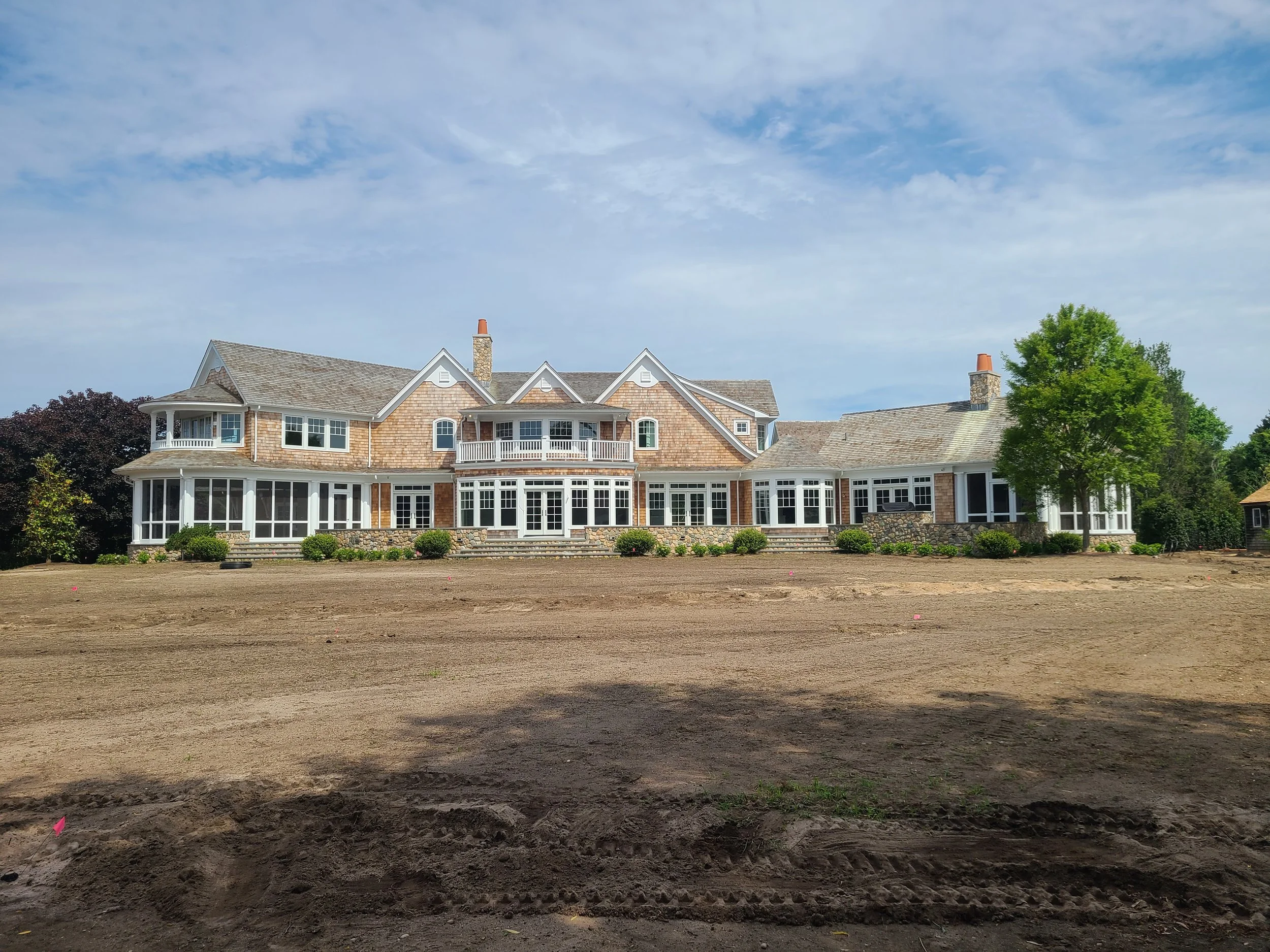 Large luxurious house with a stone and wooden exterior, multiple windows, and a spacious front yard, under a partly cloudy sky.