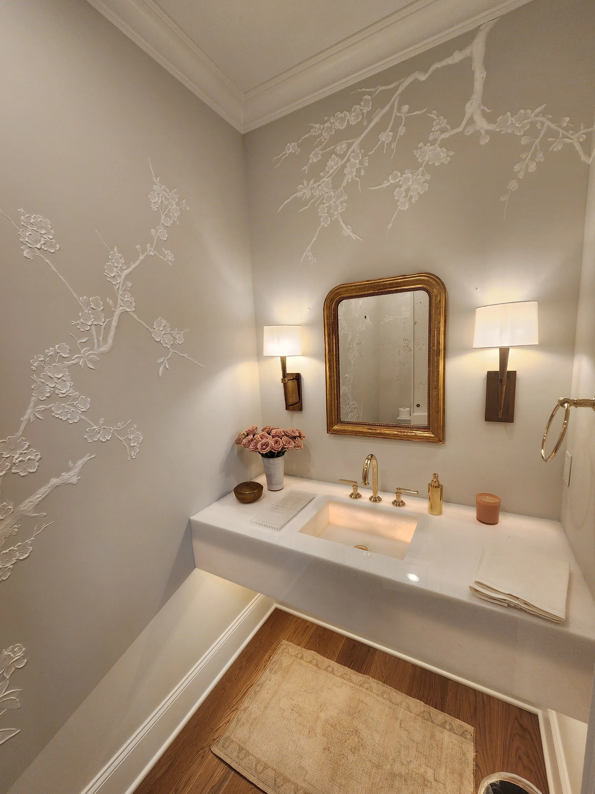 Bathroom with white textured floral wall art, gold framed mirror, two wall sconces, white countertop with sink, pink roses in a vase, gold soap dispenser, brown and pink containers, beige towel, and a beige rug on wooden floor.