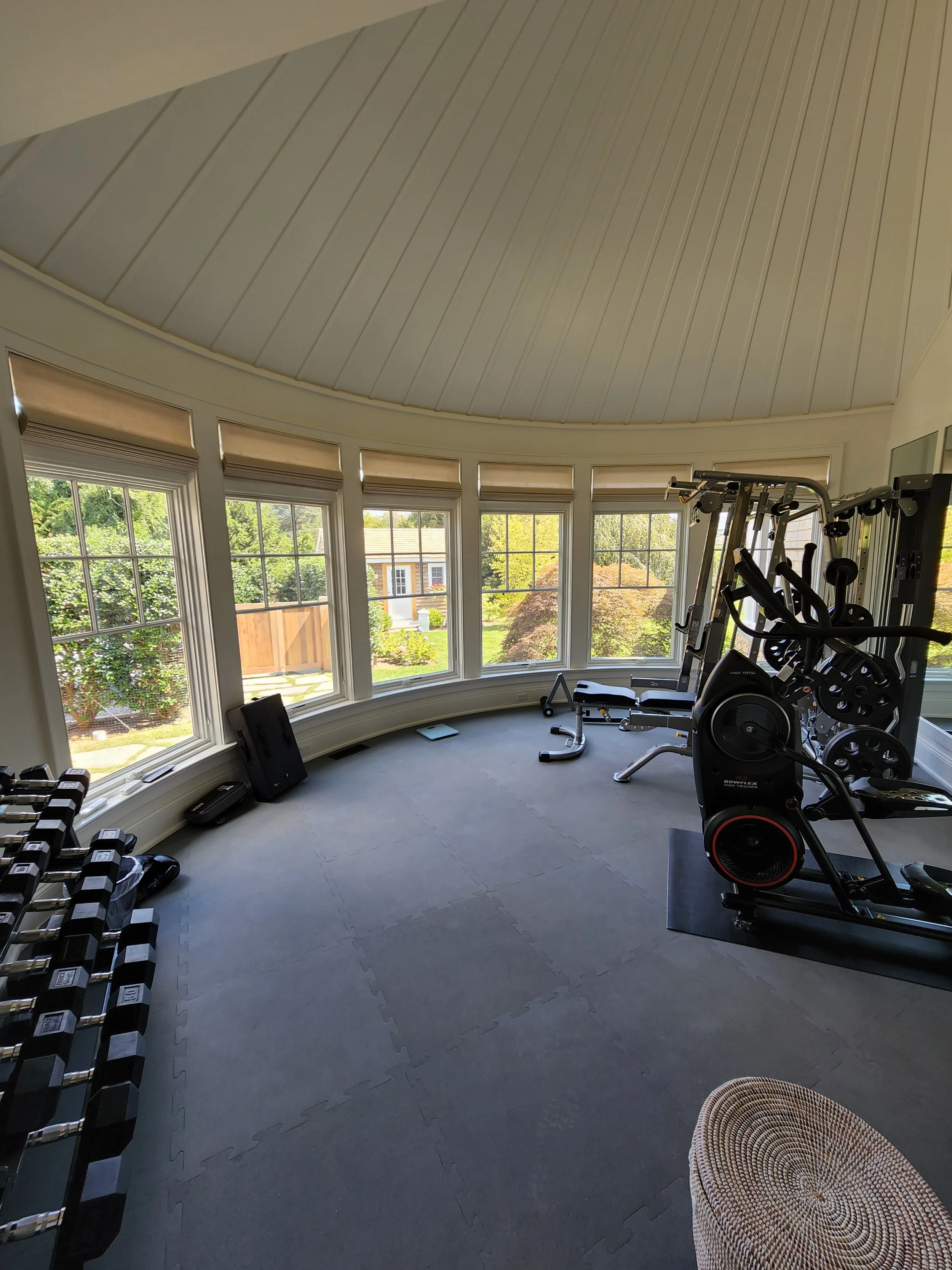 Home gym with dumbbells, elliptical machine, and sunlight through large windows revealing a green yard.