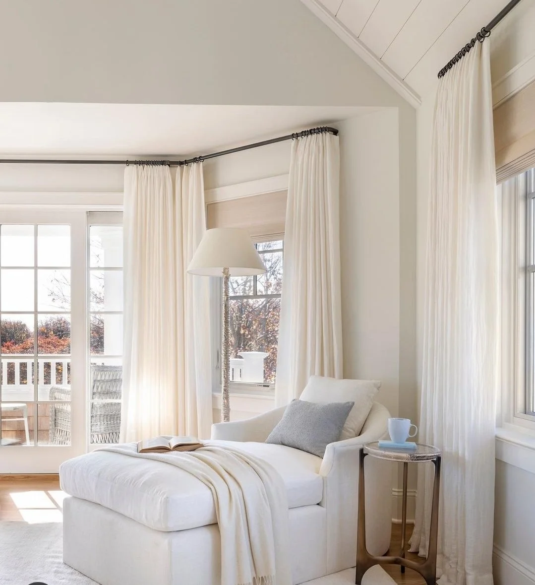 Bright living room with white chaise lounge with gray pillow, an open book, cream curtains, a floor lamp, and a small side table with a cup, near windows and a door leading to an outdoor balcony.