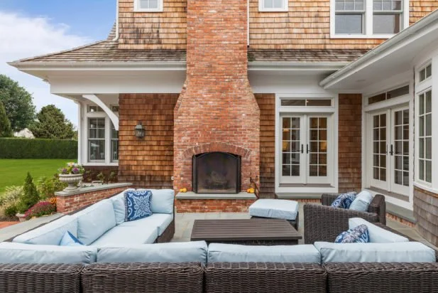 Outdoor patio with wicker furniture, light blue cushions, and throw pillows in front of a brick fireplace on a house with white and brick exterior walls.