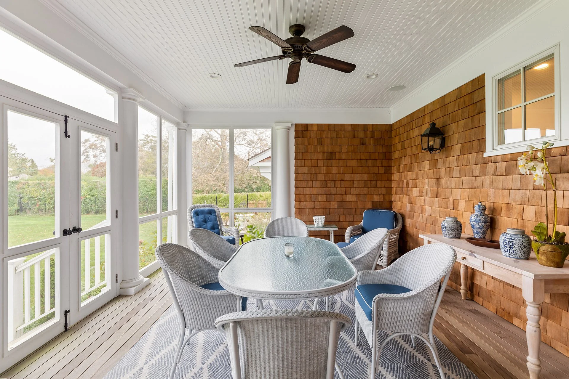 Enclosed porch with a glass-top dining table, white wicker chairs with blue cushions, a wooden sideboard with blue and white vases and a potted orchid, window and door overlooking a garden, wooden shingle accent wall, ceiling fan, and white beadboard