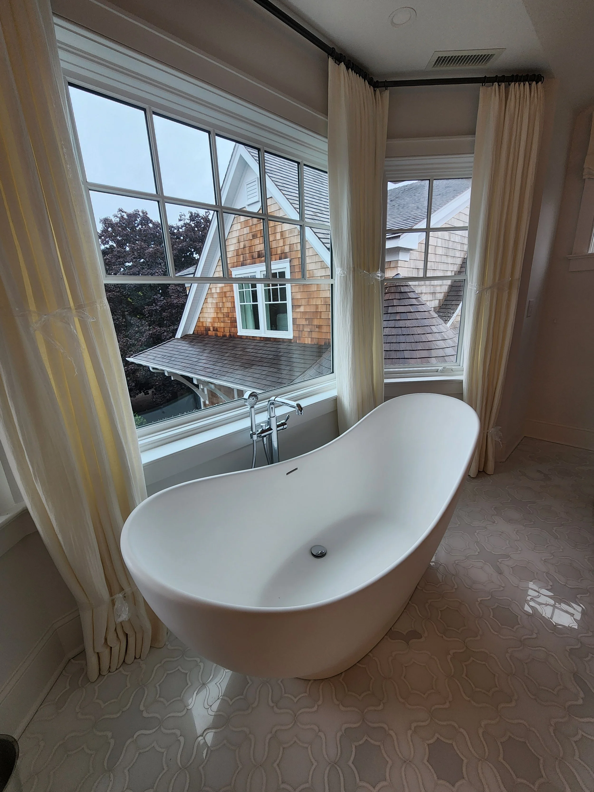 A freestanding white bathtub next to large windows with cream-colored curtains, overlooking rooftops and trees outside.
