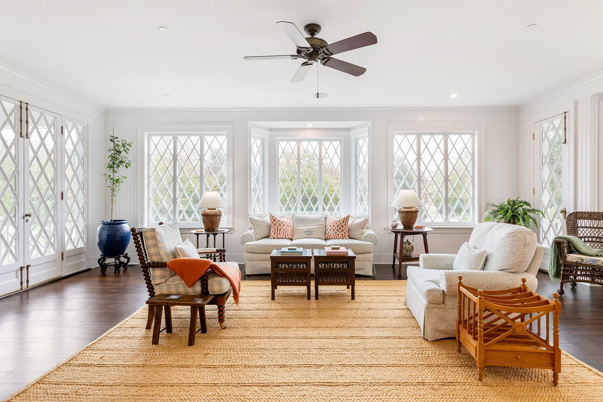 Bright living room with white panel walls, large diamond window panes, a ceiling fan, and various white and wooden furniture, including sofas, chairs, side tables, and lamps, on a woven beige rug.