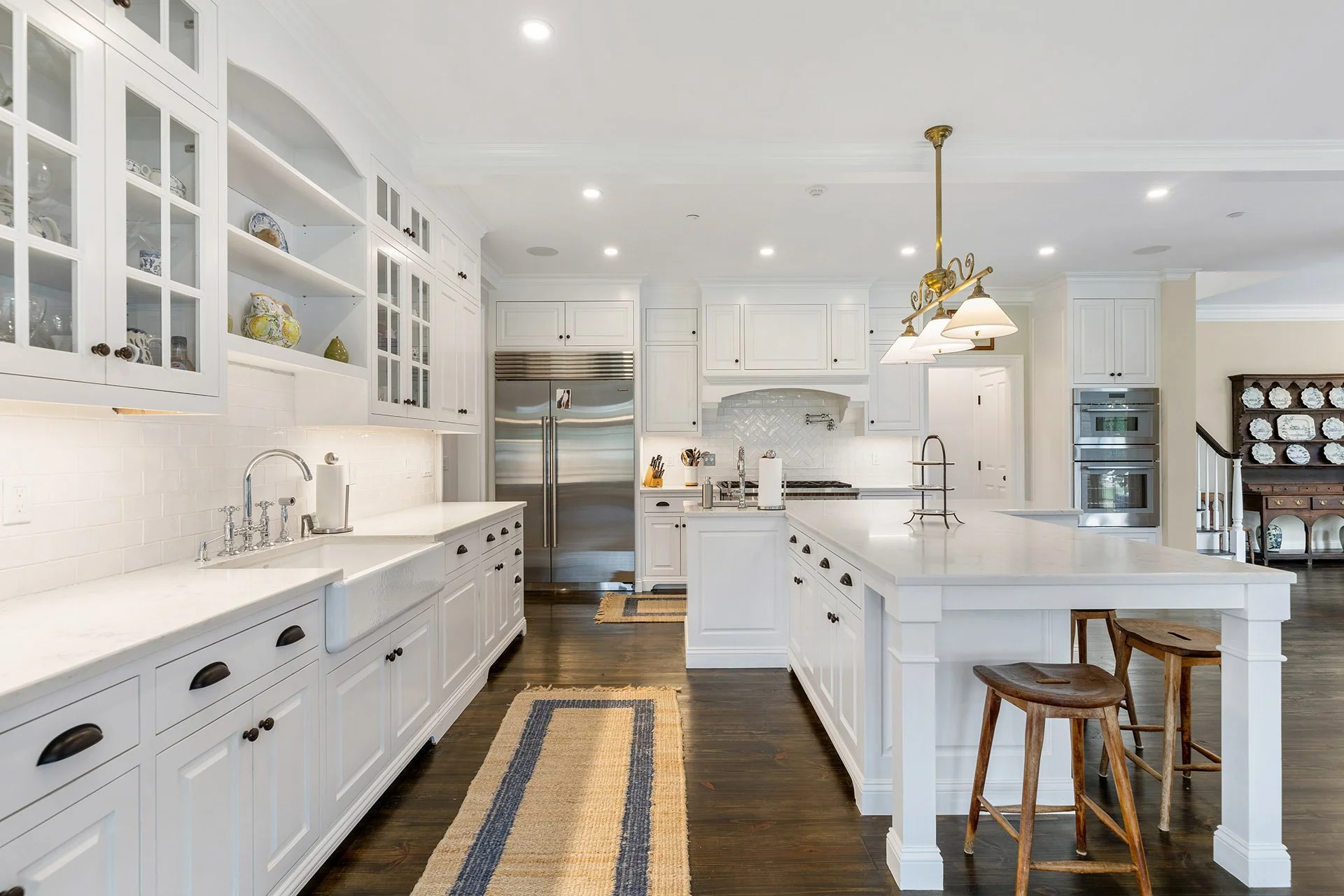 Modern white kitchen with dark hardwood floors, white cabinets, a large island with a breakfast bar, stainless steel refrigerator, and a hanging light fixture.
