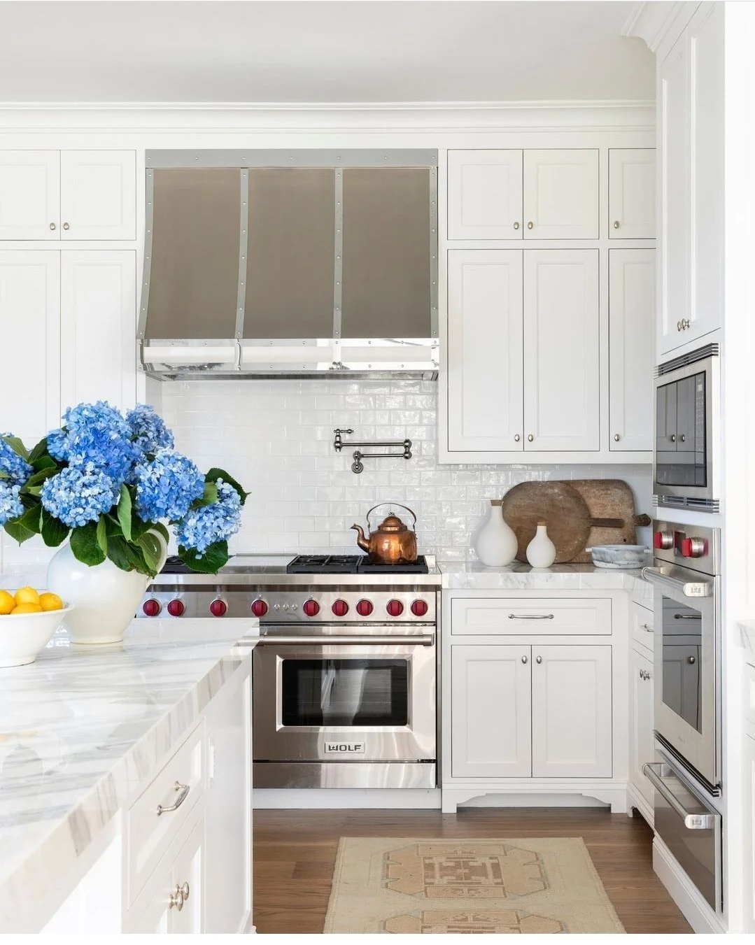 Modern kitchen with white cabinetry, stainless steel appliances, a copper teapot on the stove, and a large vase of blue hydrangeas on the counter.