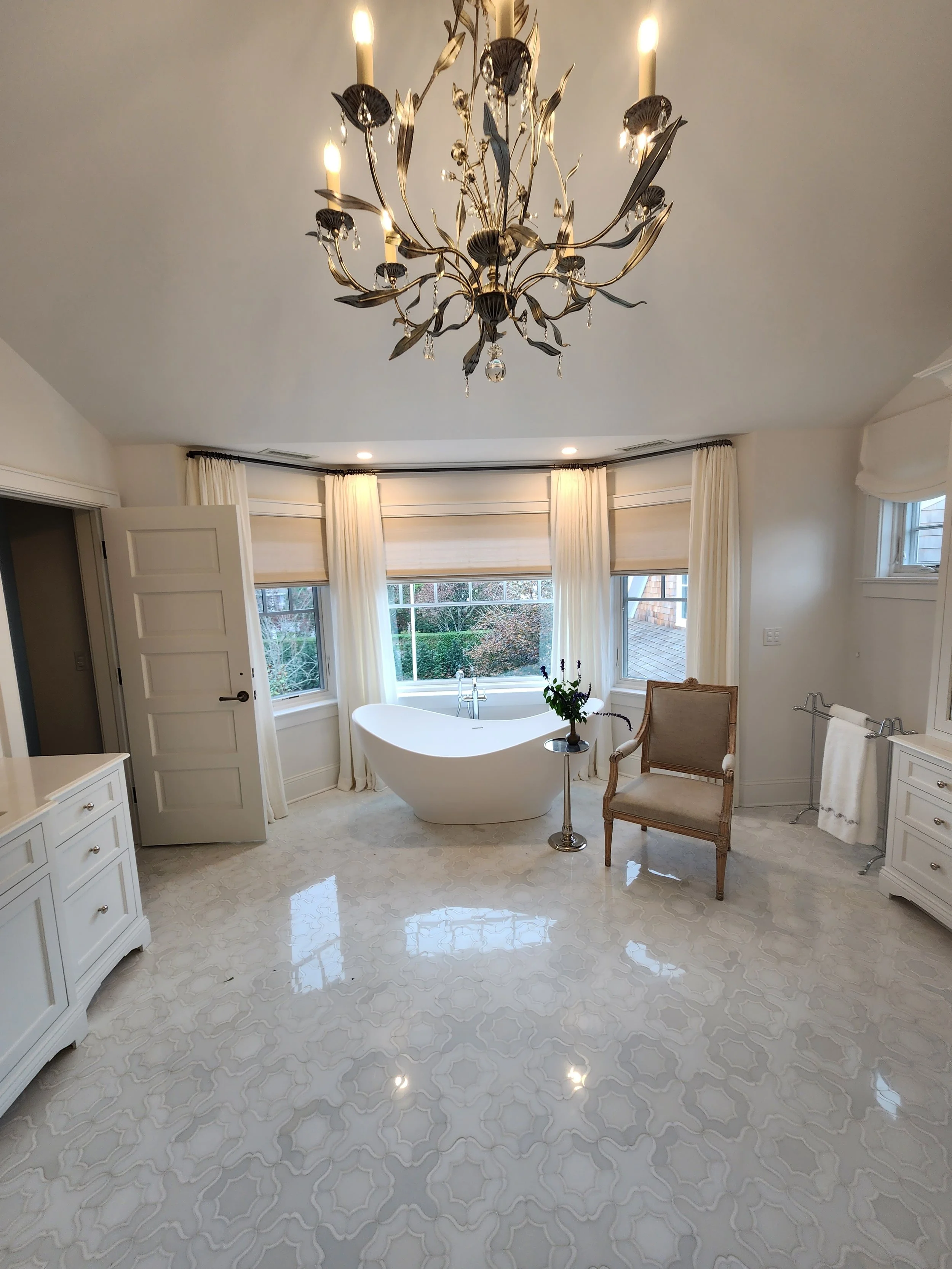 Elegant bathroom with white bathtub, chandelier, cream curtains, and patterned marble floor.