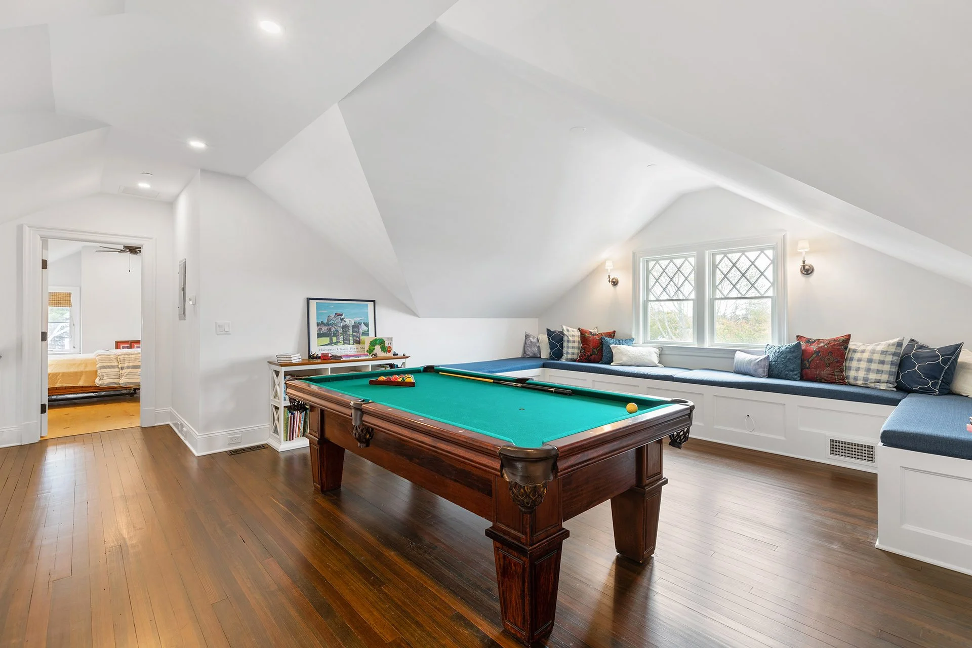 A cozy attic recreation room with a pool table, built-in window seat with pillows, hardwood floors, and white walls.