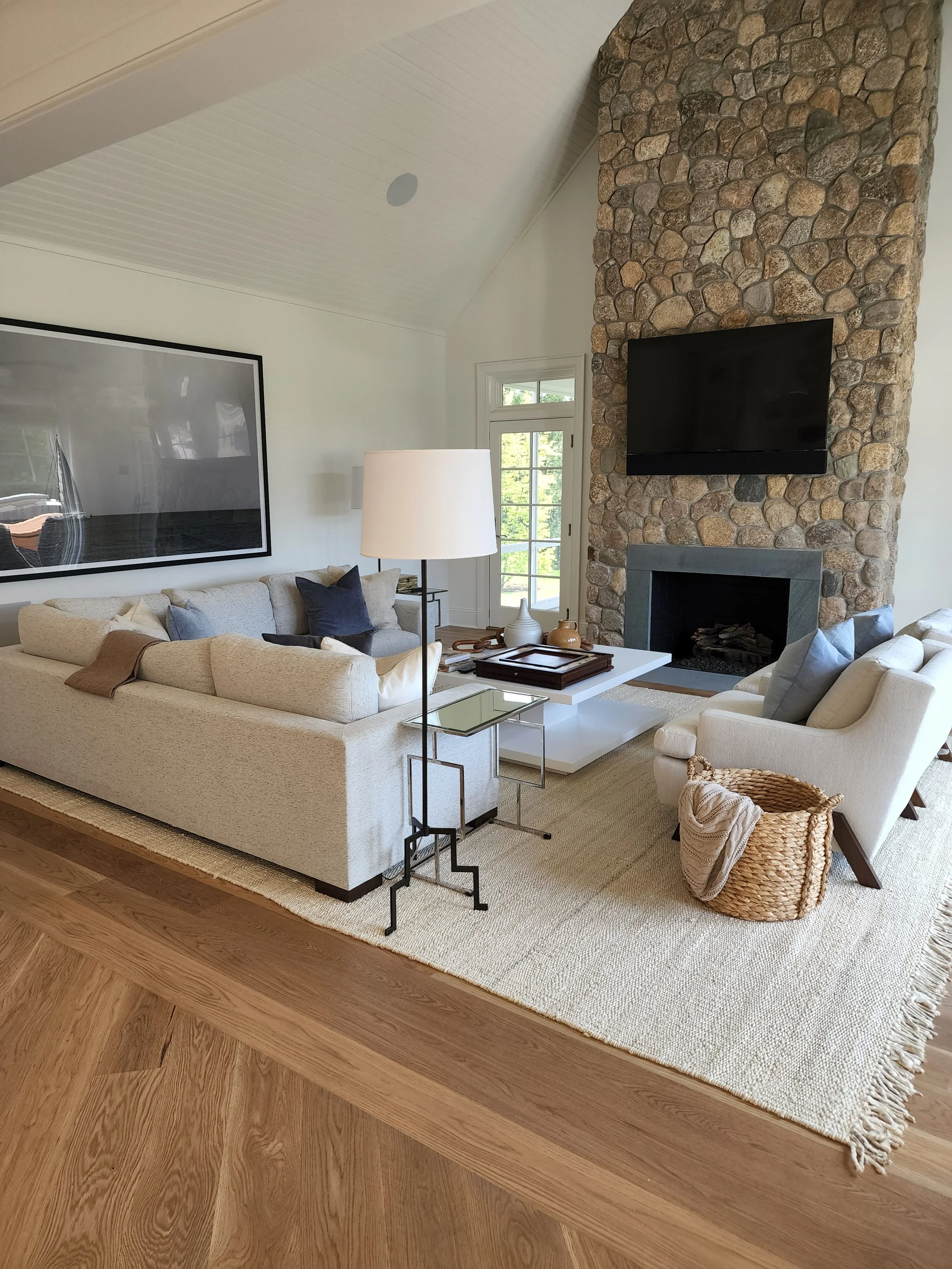 Living room with beige sofa, white armchair, stone fireplace with a mounted TV, glass coffee table, large framed artwork, floor lamp, and woven basket on a beige rug, with a window showing greenery.