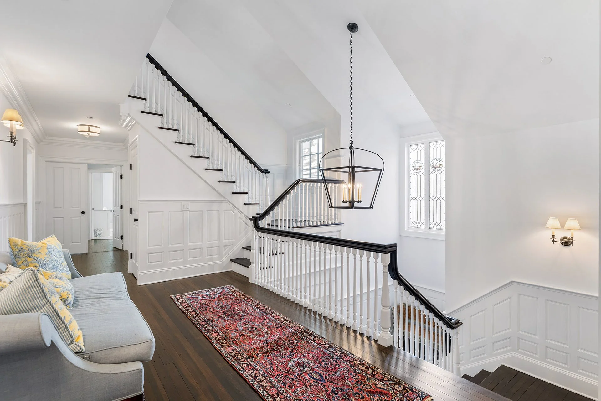 Interior view of a bright, modern staircase and hallway, with white walls, dark wood flooring, a colorful rug, a light gray sofa with patterned pillows, and multiple light fixtures including wall sconces and a hanging chandelier.