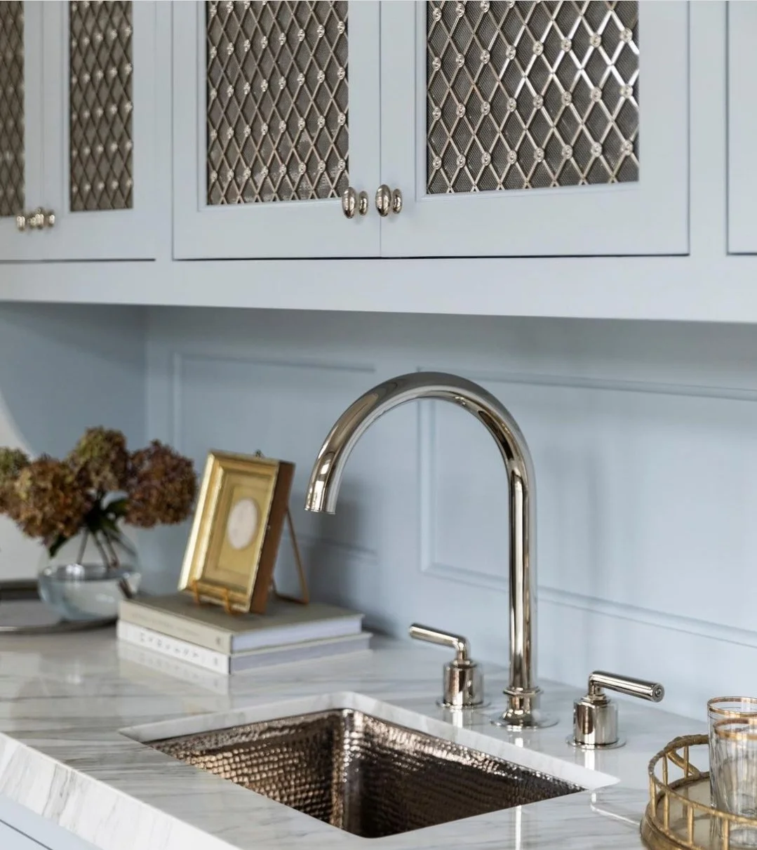 A kitchen sink with a polished metal faucet and handles, white marble countertop, and light blue cabinets with decorative lattice-style glass doors, a small picture frame, a vase of dried flowers, and a gold tray with candles.