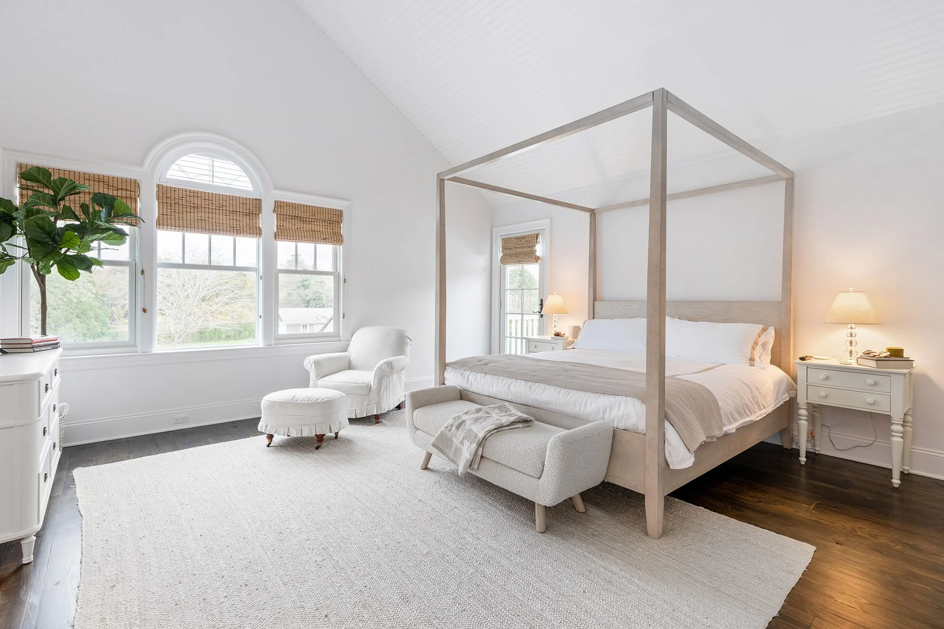 Bright bedroom with a four-poster bed, white armchair with matching ottoman, large windows with bamboo blinds, dark hardwood floor, and neutral-colored decor.