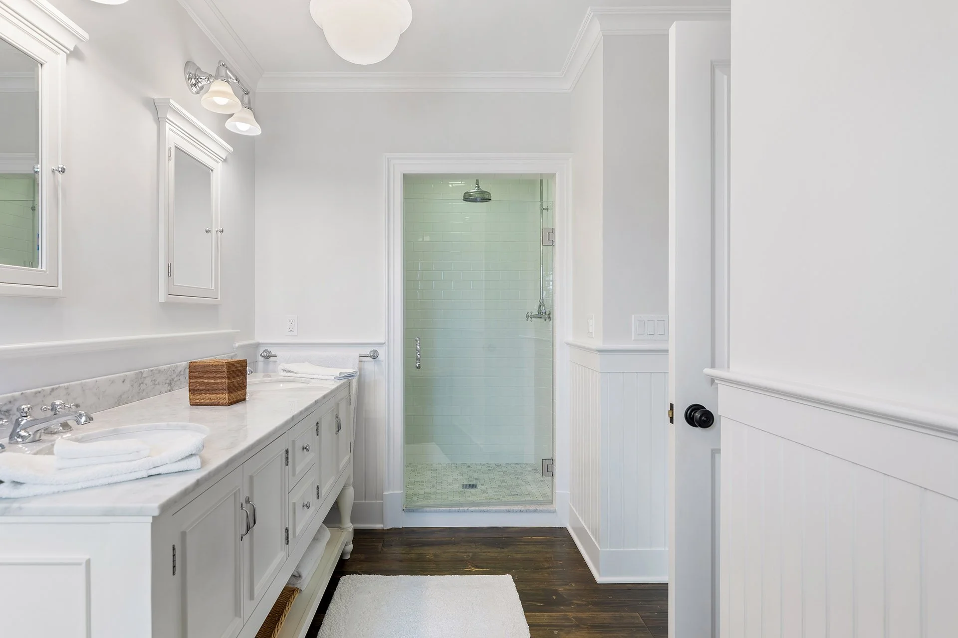 Modern white bathroom with a glass shower enclosure, white vanity with marble countertop, wall-mounted mirrors, and dark hardwood flooring.