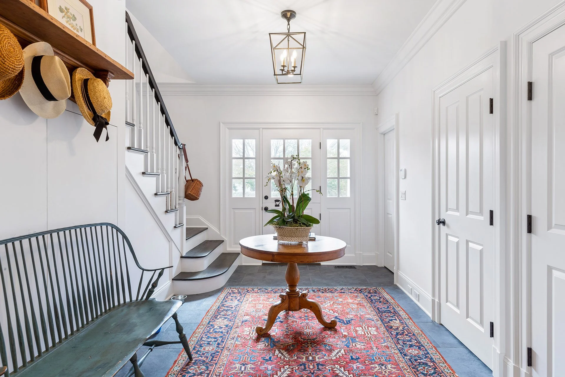 Entryway with white walls, front door with glass panes, a round wooden table with a potted orchid, a patterned rug, a black bench, and a staircase with hats hanging on the wall.