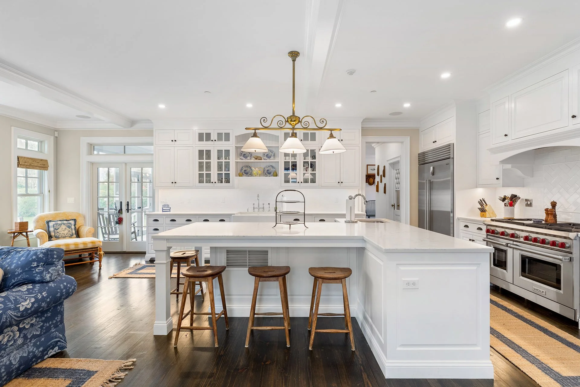 Bright white kitchen with large island, three wooden bar stools, stainless steel appliances, and a cozy sitting area with a yellow armchair and TV.