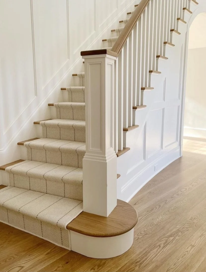 Interior staircase with a curved wooden landing, white wall paneling, beige patterned carpet on stairs, and wooden handrails.