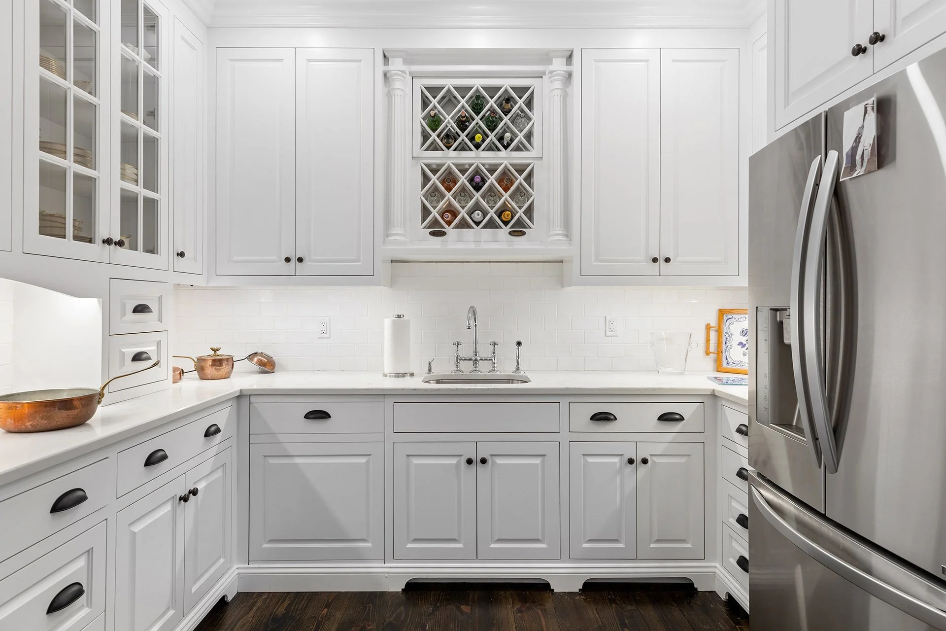 White kitchen with cabinets, a built-in wine rack with bottles, stainless steel refrigerator, and dark wooden floor.