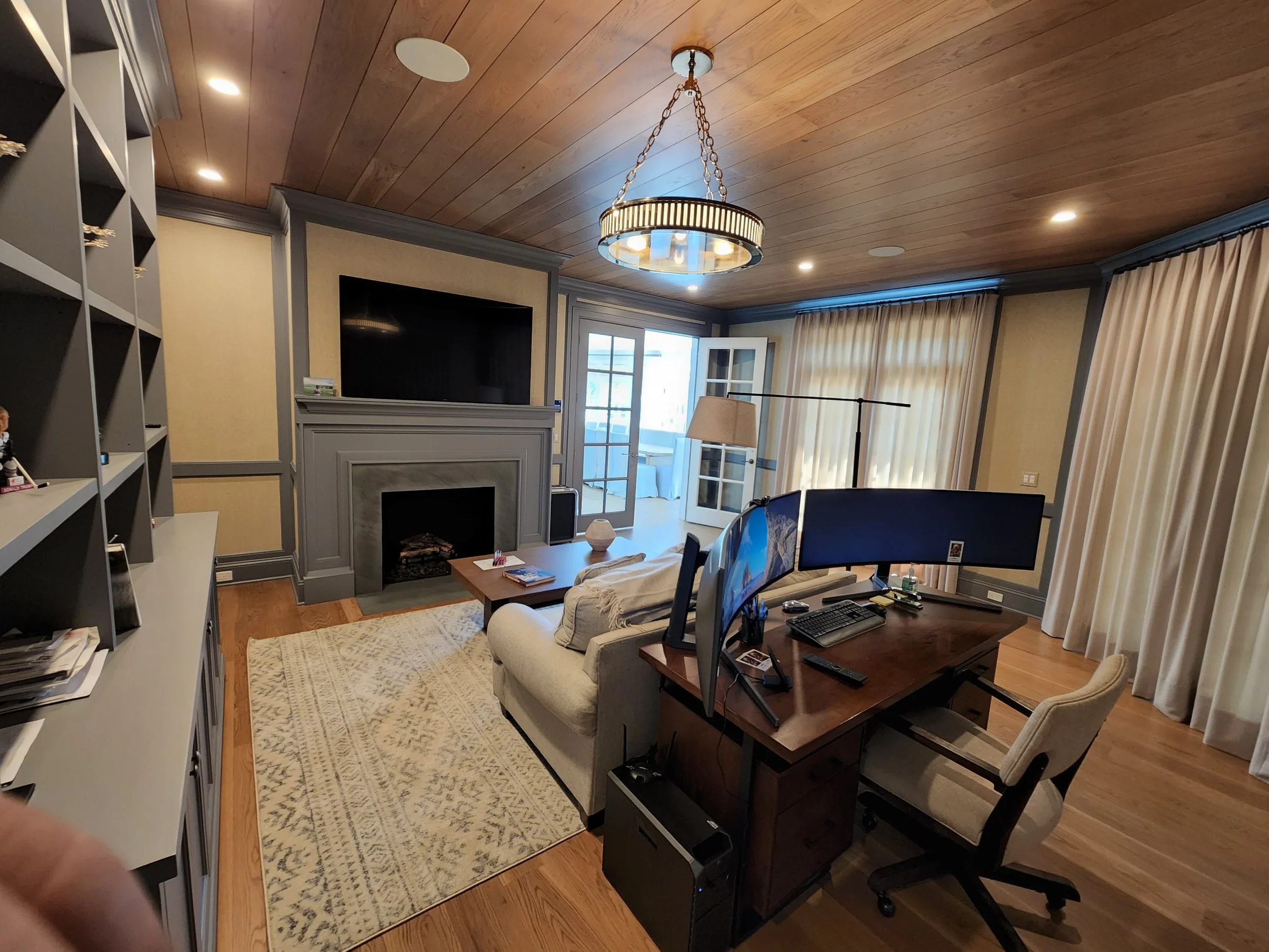 Living room with a fireplace, built-in shelves, a sofa, a coffee table, a large TV, and a desk with computer monitors, in front of curtains covering windows and glass doors.