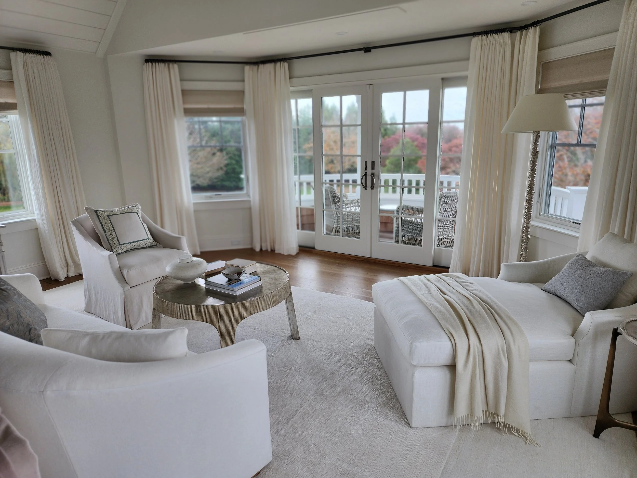Bright living room with white furniture, large windows with cream curtains, and a balcony with outdoor seating, overlooking trees with fall foliage.