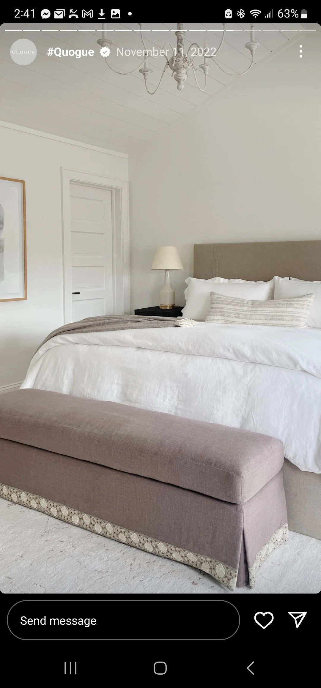 A neatly made bed with white bedding and pillows, a bedside table with a lamp, a beige upholstered headboard, an upholstered bench at the foot of the bed, a door, and a chandelier in a bright, minimalist bedroom with white walls and trim.