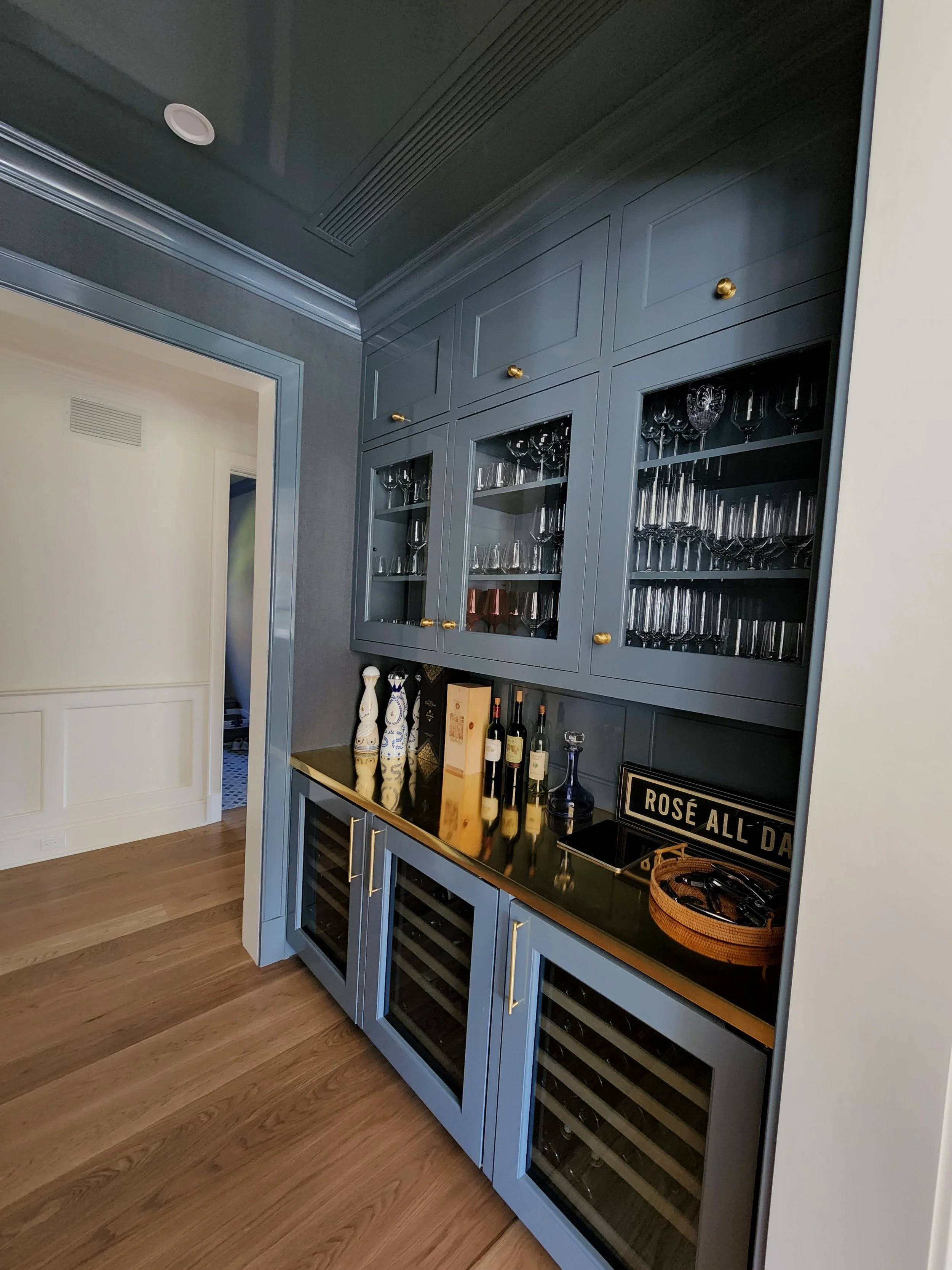 Built-in gray cabinet with glass doors displaying wine glasses and bottles, a black countertop with decorative vases, wine bottles, a framed sign, and a basket, against a wooden floor in a dining or bar area.