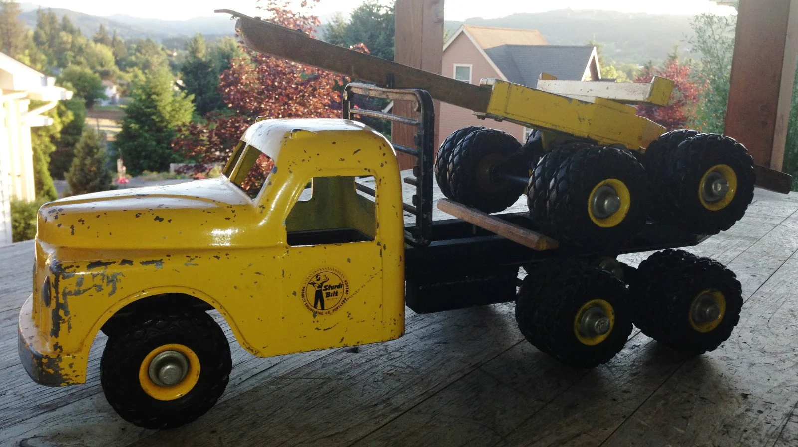 Yellow toy dump truck with black wheels and a red truck bed, sitting on a wooden surface with a scenic background of trees and houses.