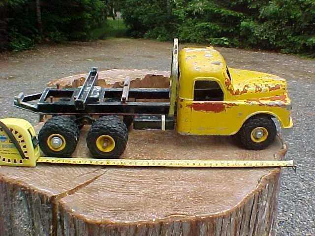 A vintage yellow toy truck with a flatbed, placed on a tree stump, with a tape measure stretched across the stump.