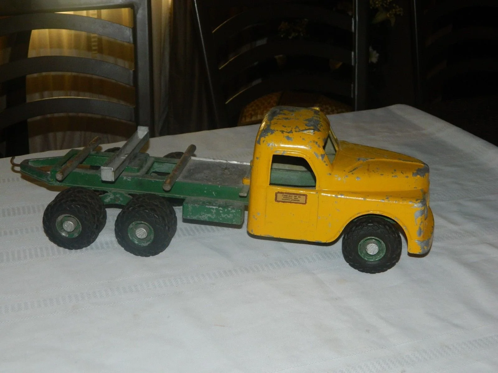 A vintage toy tow truck with a yellow cab and green flatbed, showing chipped paint, on a white tablecloth with black chairs in the background.