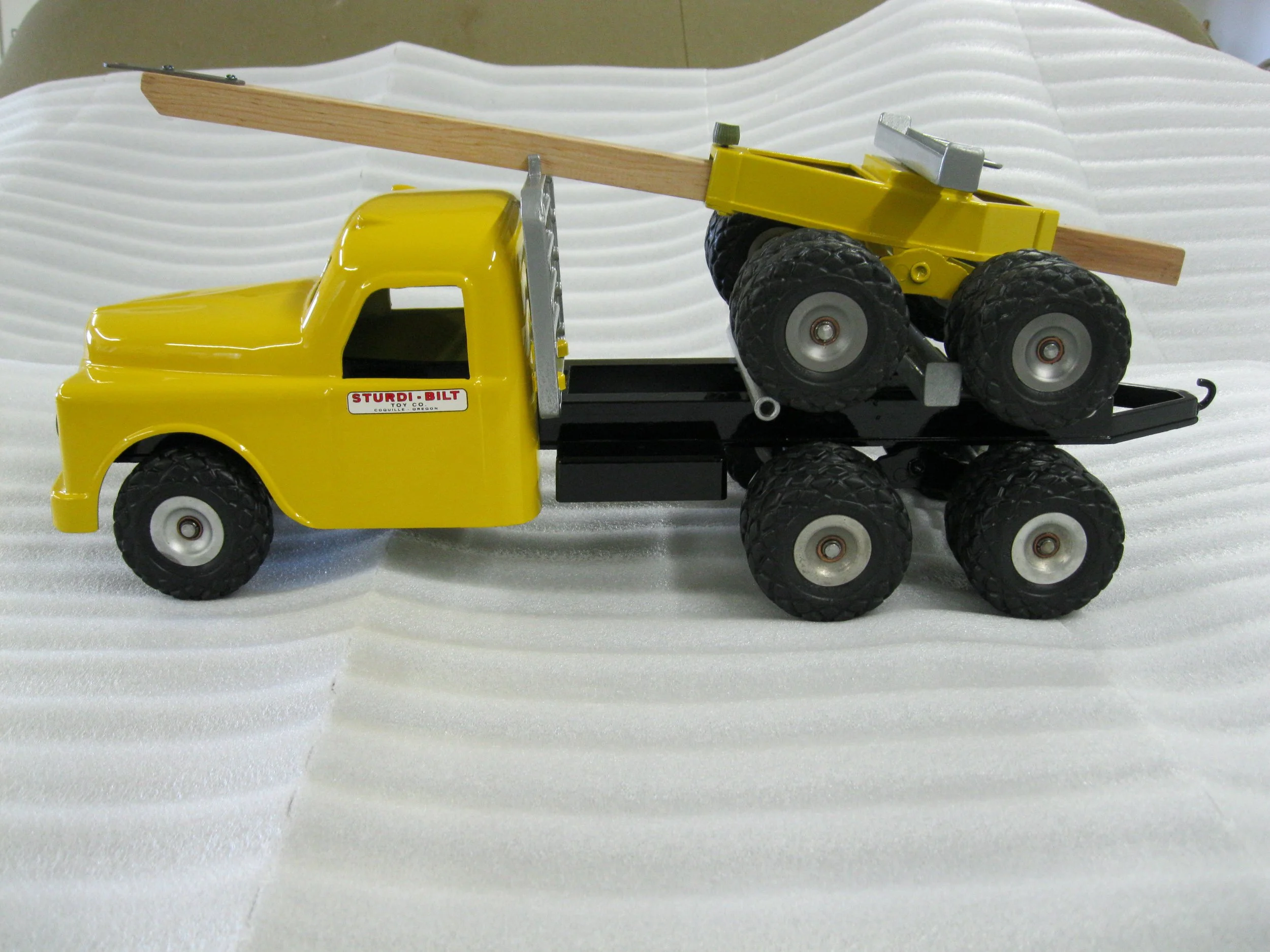 Yellow toy truck with six black wheels, carrying a small yellow toy bulldozer with a wooden blade.