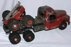 A vintage red toy dump truck with a trailer carrying a toy cement mixer, both showing signs of wear and rust.