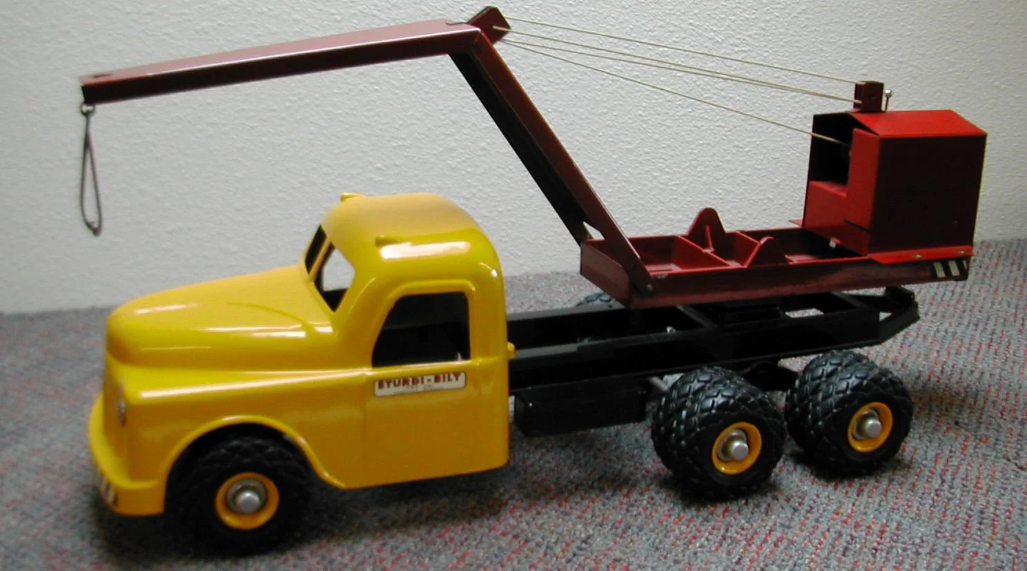 A toy tow truck with a yellow cab, black chassis, and red towing arm and storage box, positioned on a carpeted floor against a white wall.