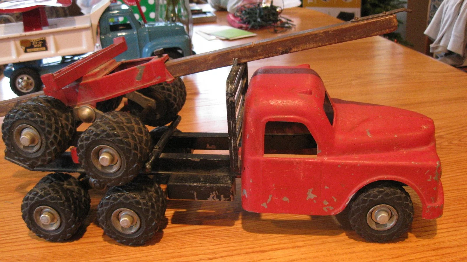 A vintage red toy truck with a flatbed carrying six small tires and a wooden plank is placed on a wooden surface. The truck has chipping paint and a simple design.
