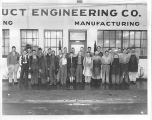 Group photo of factory workers standing outside a building with signs 'Engineering Co.' and 'Manufacturing.'