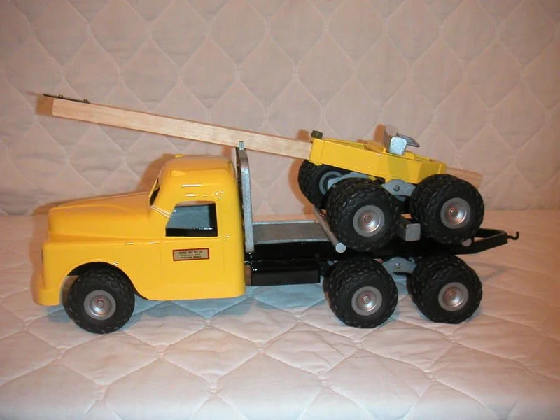 A yellow toy tow truck with a yellow toy bulldozer attached to its flatbed, placed on a quilted surface with a patterned background.