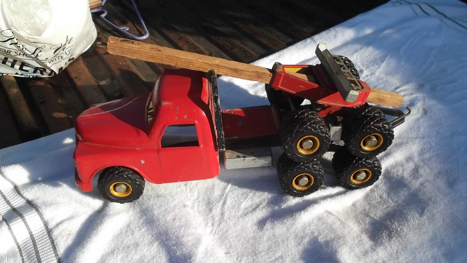 A vintage red toy truck with six large black wheels is on a white cloth outdoors, with a piece of wood resting across its bed, and a wooden surface and other objects in the background.