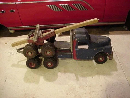 A vintage toy tow truck with a crane arm, parked on a garage floor next to a red vehicle.