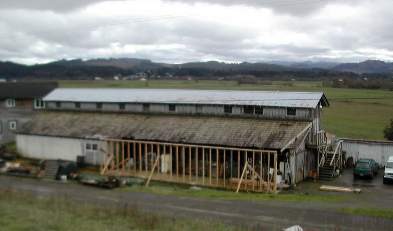 An old wooden barn with a weathered metal roof and partially under construction, surrounded by parked cars and construction materials, with a rural landscape and cloudy sky in the background.