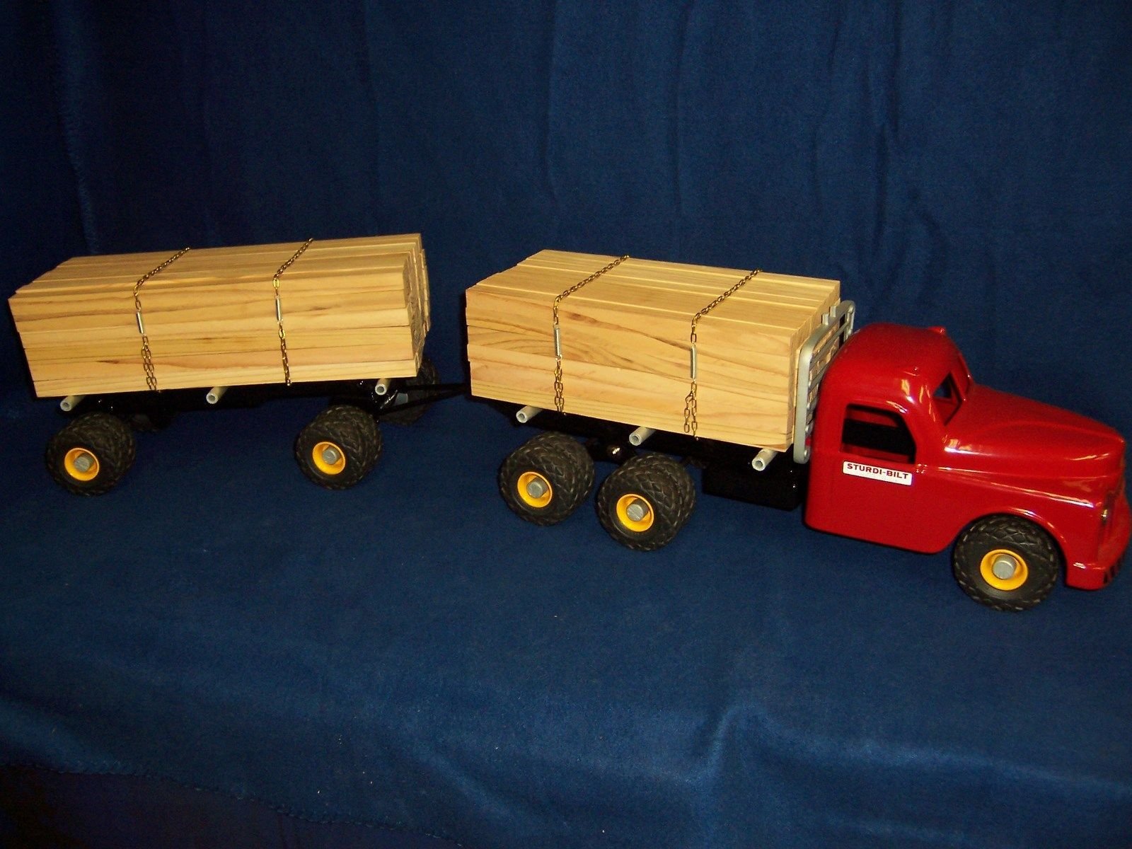 Toy red truck with two flatbeds carrying miniature wooden planks, set against a dark blue background.