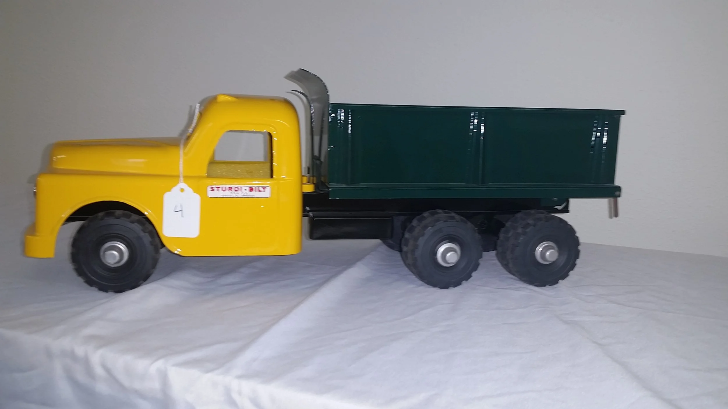 Yellow toy dump truck with black wheels and green bed, placed on a white surface against a plain wall.