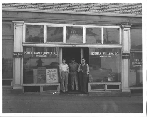 Three men standing in front of a storefront with signs for Power Brake Equipment Co. and Norman Williams Co.