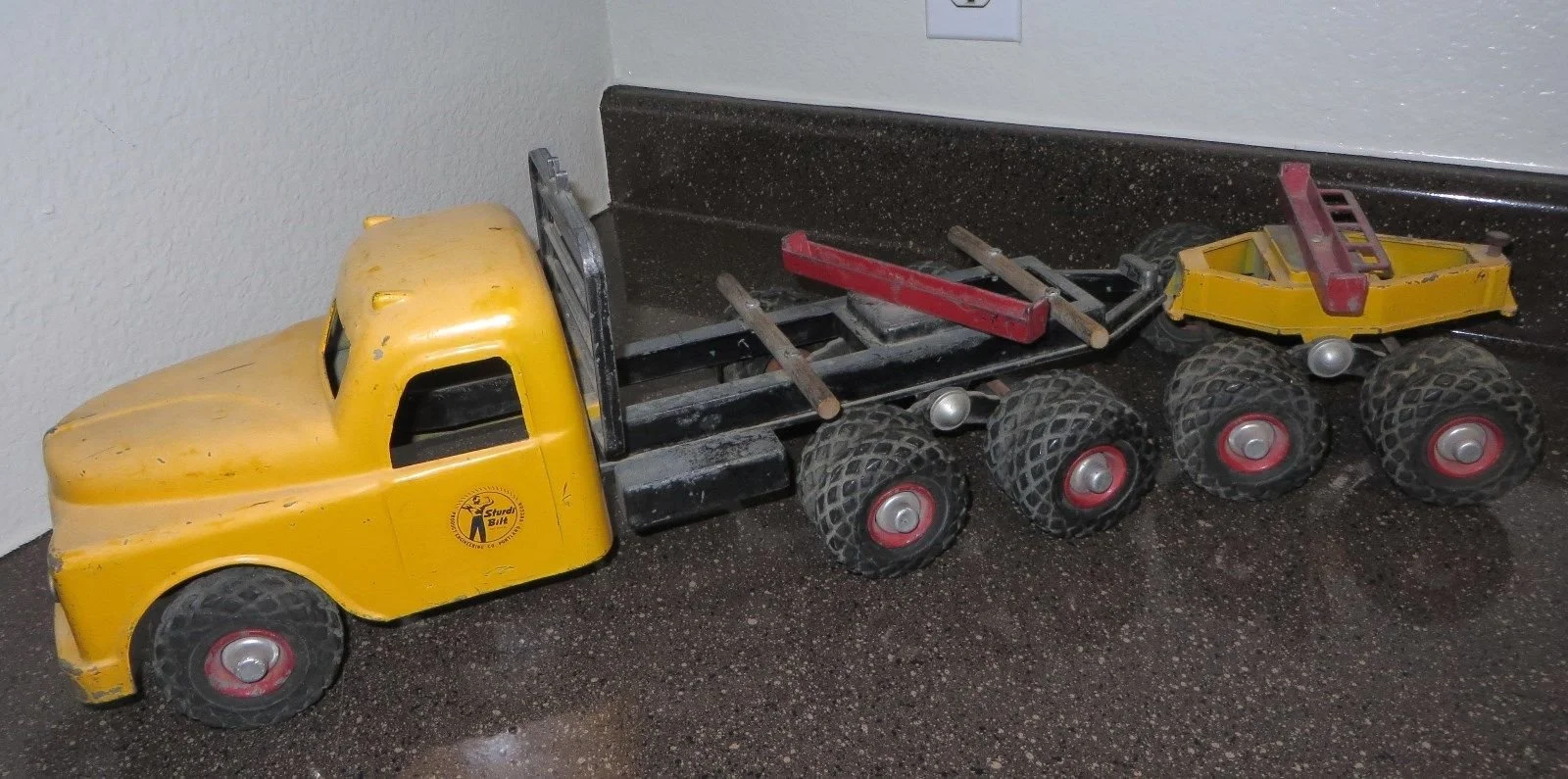 A toy yellow pickup truck with six wheels is placed on a speckled dark countertop against a white wall.