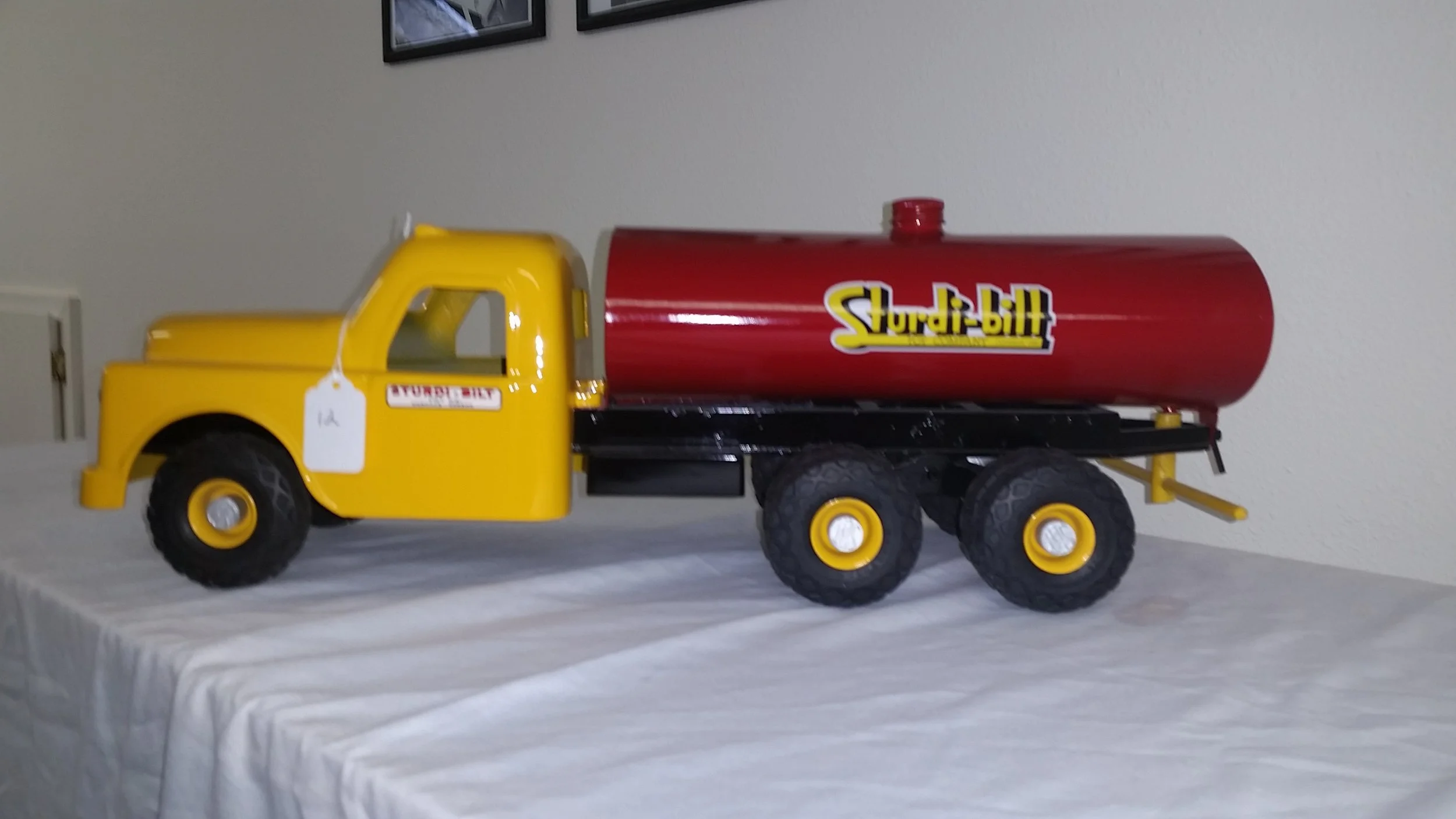 Yellow toy truck with black tires and a red tank labeled 'Sturdi-bilt' on a white table against a plain wall.