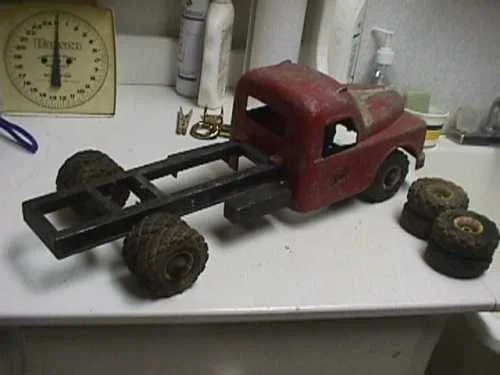 A vintage red toy truck with four wheels, missing its cargo bed, placed on a white surface with four additional wheels nearby. In the background, there is a kitchen scale and some kitchen items.