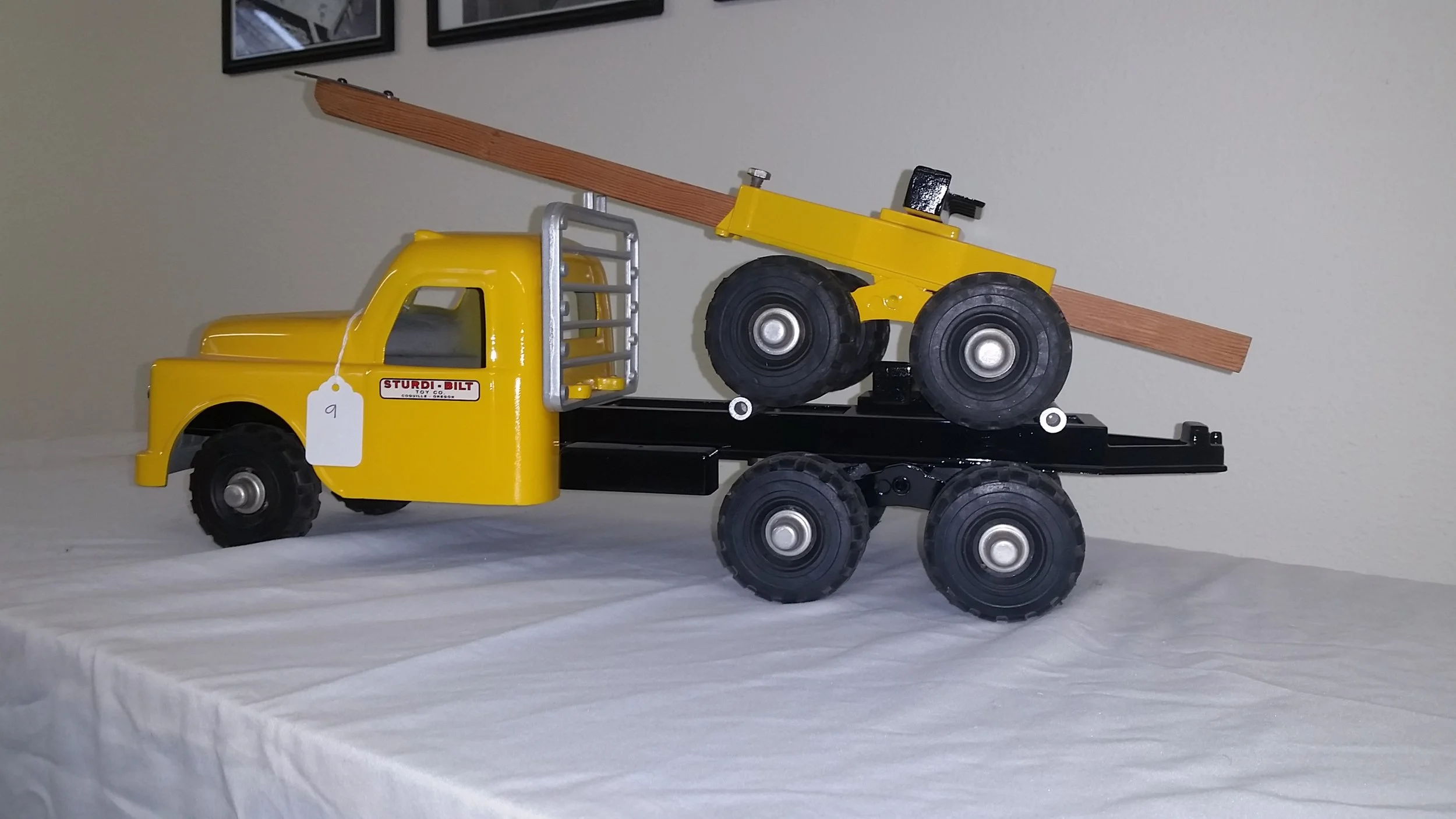 A yellow toy tow truck with black wheels and a crane arm carrying a yellow toy bulldozer with black wheels, placed on a white table against a beige wall.