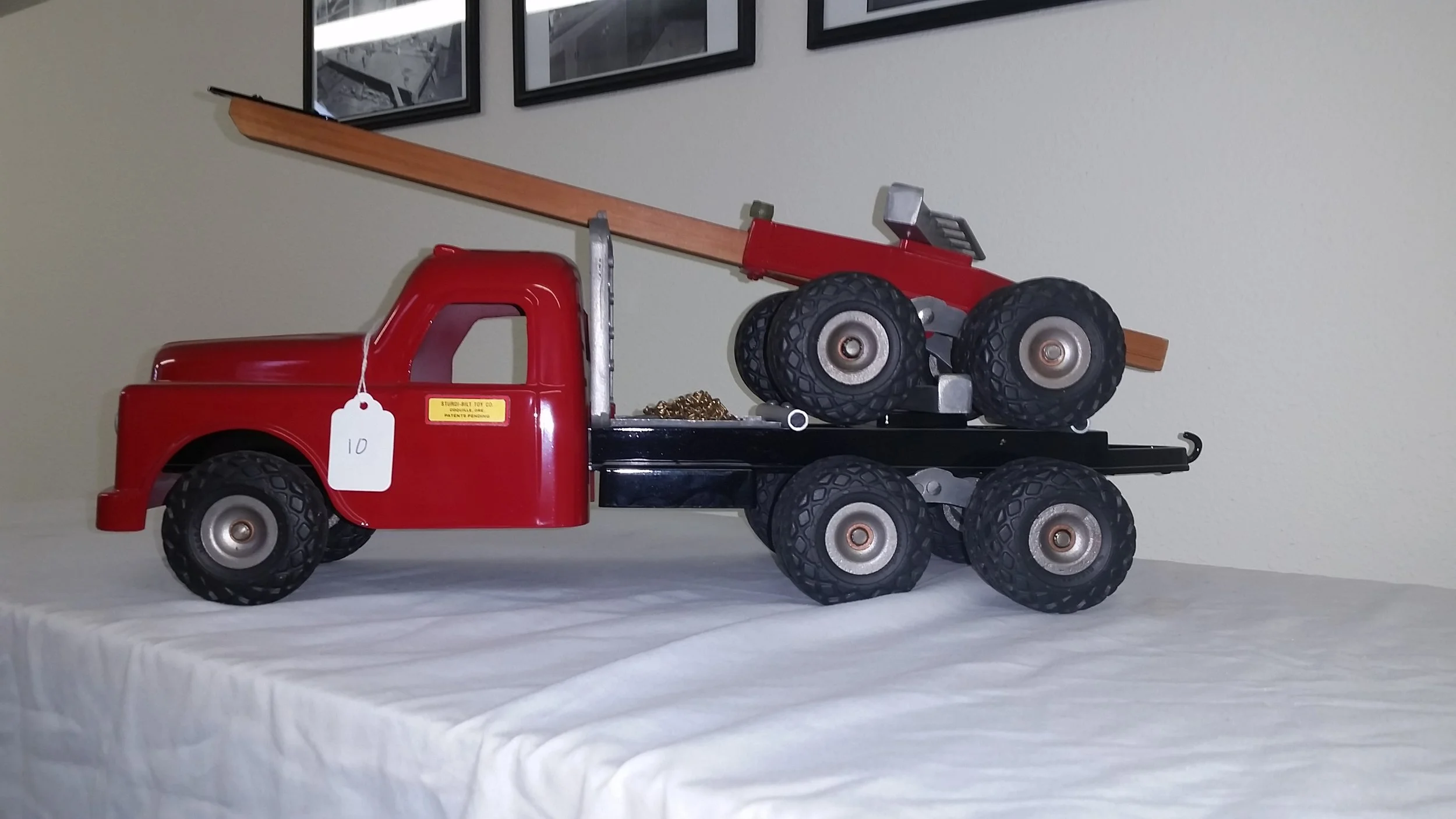 A vintage red toy tow truck with black chassis and rubber wheels, displaying a small red and gray toy car on its flatbed, with a long wooden ramp attached.